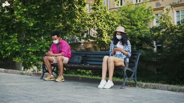 Afroamerican Man in Protective Mask Sitting on Bench of Park Using Smartphone