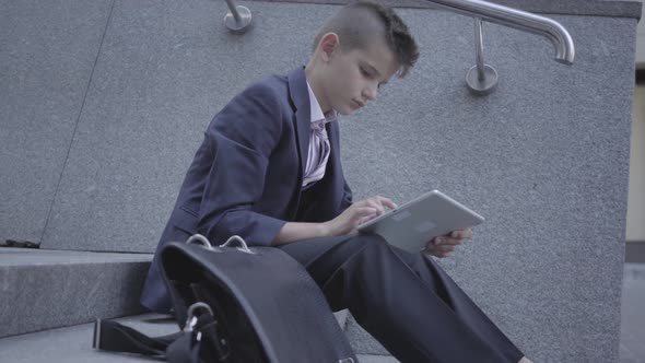 Little Boy Wearing Business Suit Sitting on the Stairs on the Street Typing on the Tablet