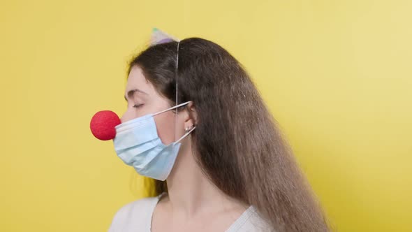 A Young Woman in a Protective Mask with a False Red Nose and a Paper Cap Turns and Looks Sad