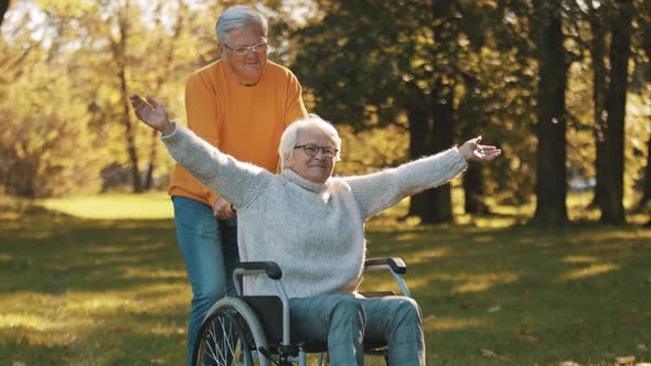 Elderly Couple in the Park. Husband Pushing His Disabled Wife with Outstretched Hands in the