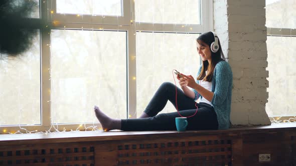 Cute Girl Brunette Is Sitting on Window Sill, Listening To Music in Headphones and Using Smartphone