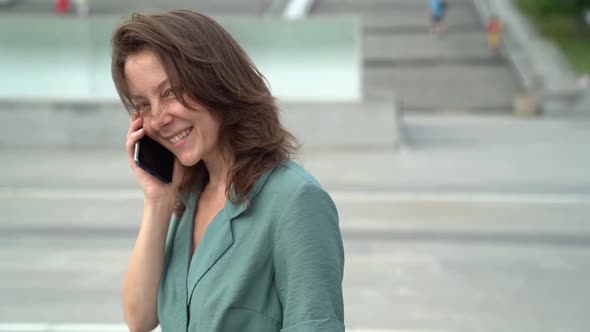 Charming Middle-aged Woman Is Chatting By Cellphone on City Street at Windy Day, Smiling To Camera