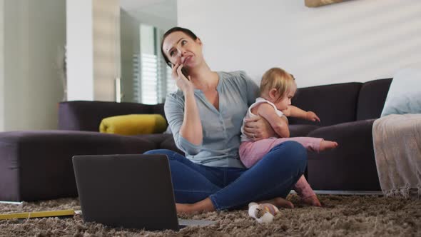 Caucasian mother holding her baby talking on smartphone while working from home