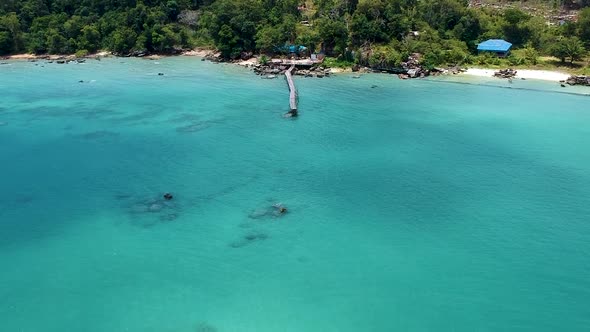 Aerial tilt reveal of old wooden pier and green coastline with ocean rocks and blue sea. Koh Rong Sa