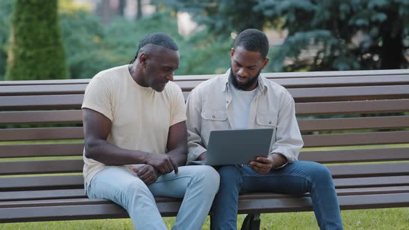Two Black Men in Casual Clothes Sitting on Bench in City Park Using Laptop