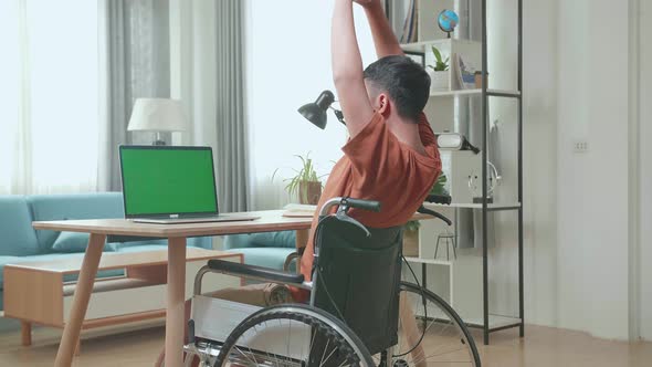 Young Asian Boy Sitting In A Wheelchair Stretching While Using Laptop With Green Screen At Home