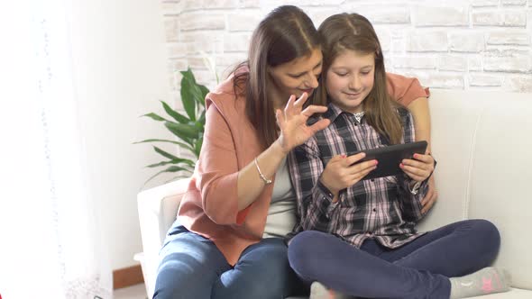 Happy mother and her daughter smiling having fun using digital tablet sitting on white sofa at home