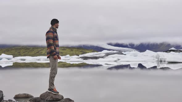 Tourist By Rocks on the Side of Frozen Lake with Melting Glacier in Background