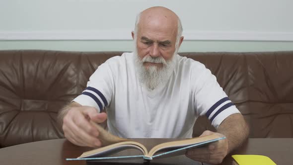 Thoughtful Bearded Adult Man Sitting at the Wooden Table on Leather Sofa Reading the Book. Education