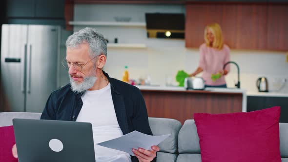 Modern Adult Graybearded Man Working with Laptop While Sitting on Sofa in Studio Kitchen