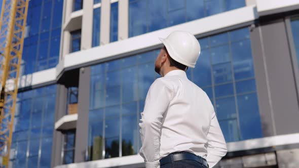 An Architect Engineer in a White Shirt and Helmet Stands with His Back to the Camera Against the