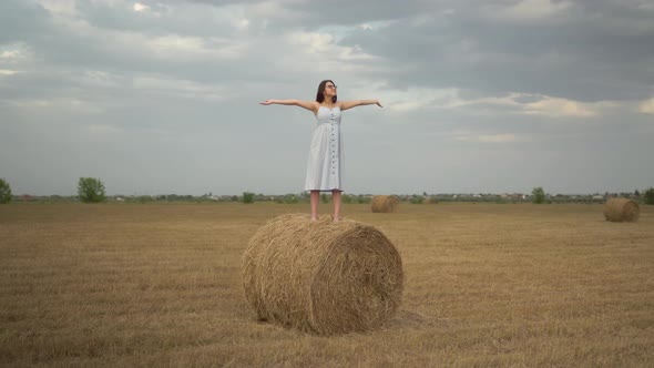 A Young Woman Stands on a Haystack in a Field and Spread Her Arms