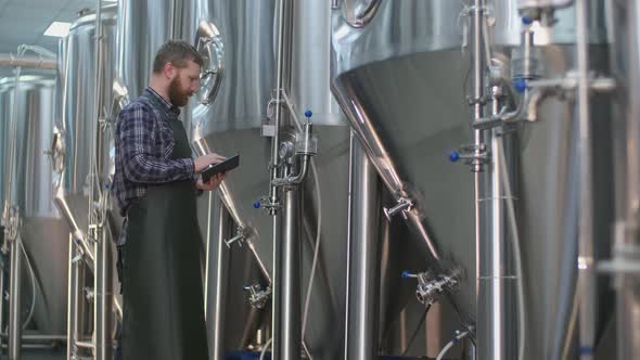 Brutal Male Brewer with a Beard Controls the Readings of Devices on Beer Tanks Using a Tablet. Craft