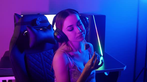 Young Blonde Gamer Woman Sits in the Chair in Neon Gaming Club and Sucking a Lollipop