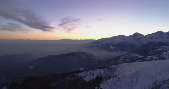 Backward Aerial Top View Over Winter Snowy Mountain and Woods Forest at Sunset or Sunrise