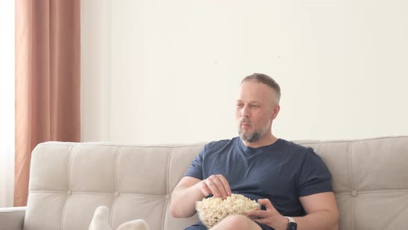 The Man is Relaxing at Home on the Couch He is Eating Popcorn and Watching TV