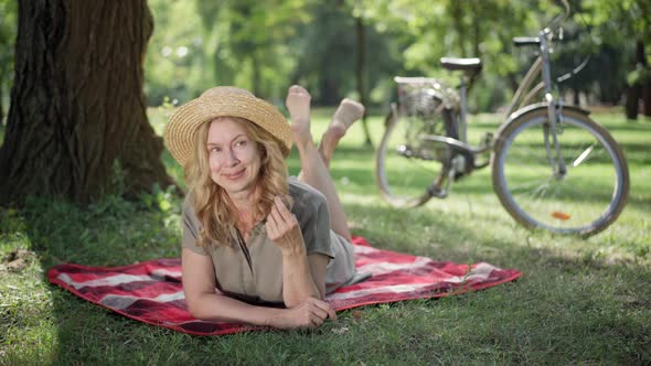 Happy Relaxed Caucasian Woman in Straw Hat Lying on Blanket in Sunny Summer Park Looking Around and