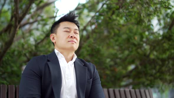 Male asian businessman relax, meditates outdoors sitting on a bench on urban