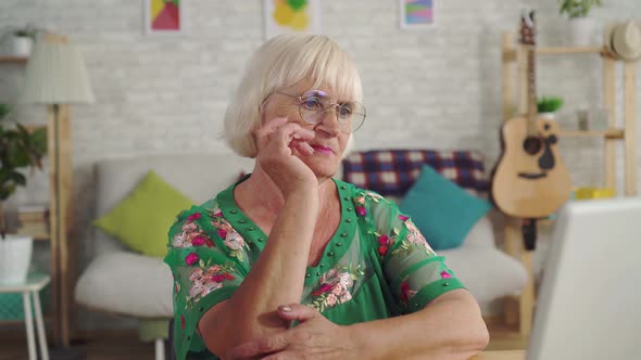 Thoughtful Old Woman Uses a Laptop While Sitting in the Living Room