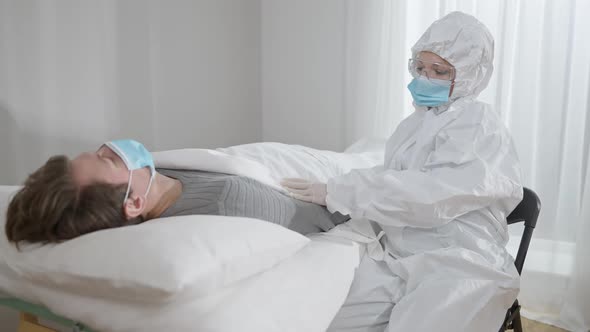 Exhausted Woman in Protective Antiviral Suit Eyeglasses and Covid19 Face Mask Holding Ill Man Hand