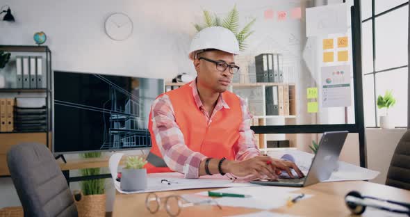 Male Engineer in Helmet and Vest Working with Computer Project and Paper Plan