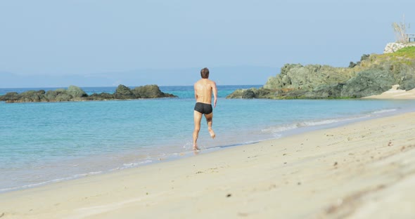 The Handsome Man with a Perfect Athletic Body in Swimming Trunks Having Fun on a Deserted Beach in