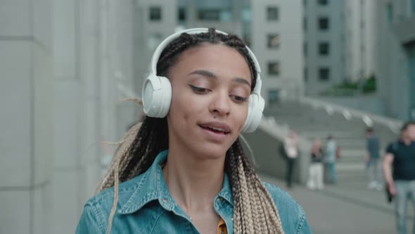 A Young African American Girl in Headphones is Listening to Music and Amusingly Dancing on a City