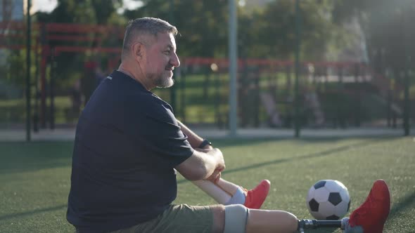 Side View Happy Bearded Amputee Sportsman Talking Sitting on Summer Sports Field in Sunlight