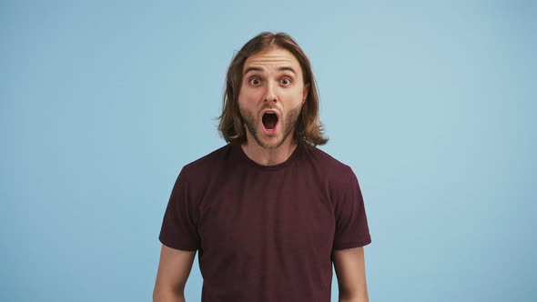 Young Bearded Man in Brown Tshirt is Saying Wow Smiling and Looking Surprised While Posing on Blue