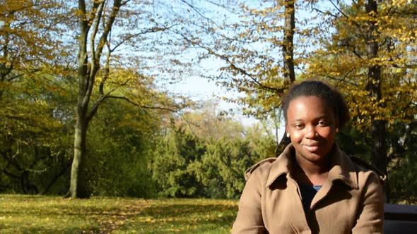 Young Beautiful African Happy Girl Sits on Bench in Woods and Stares To Camera