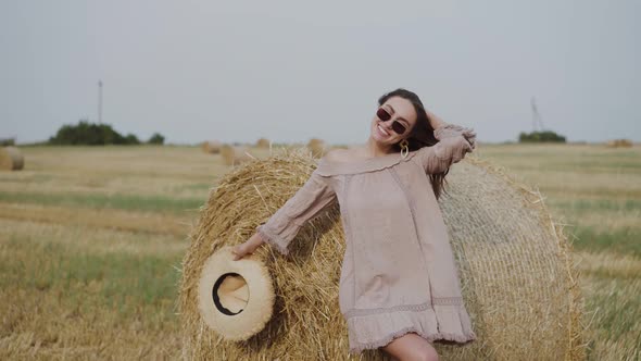 Plauful Woman Rejoices at Haystack When Holds Hat and Corrects Hair During Wind