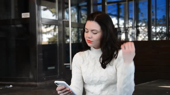 Attractive Young Brunette Woman Portrait in a White Springe Coat with Red Lips in the City Tapping