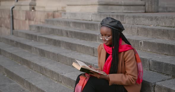 A Young Black Girl is Sitting on the Stairs on the Street and Reading a Book