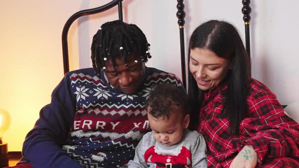 Portrait of Young Multiracial Parents Sitting on Bed with Toddler Holding a Smartphone