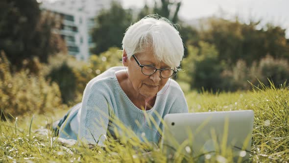 Senior Retired Gray Haired Woman Using Laptop in the Nature While Lying on the Grass