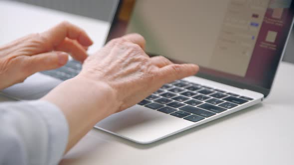 Old woman hands type on black keyboard of grey laptop on white table