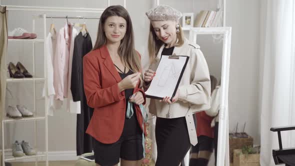 Two Beautiful Fashion Designers Showing Dress Design Sketch and Talking at Camera