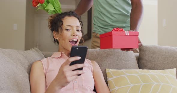 Surprised biracial woman sitting on sofa with smartphone getting present from male partner