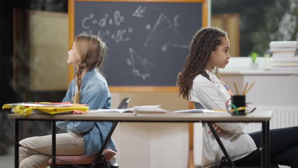 Argued Schoolgirls Sitting Back to Back in Classroom Talking and Showing Tongue Out