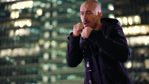 a Bald Unshaven Man is Boxing Against the Background of Blurred Highrise Buildings and City Lights