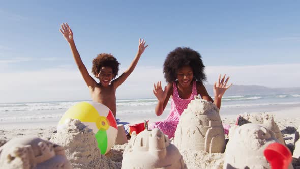 Smiling african american brother and sister with arms in the air, playing with sand on the beach