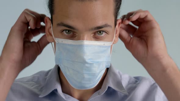 Happy Adult Male with Hazel Eyes Wearing Collared Button Down Shirt Puts on Surgical Mask on a Gray