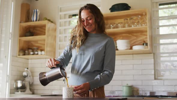 Happy caucasian woman standing in cottage kitchen pouring coffee from pot and smiling