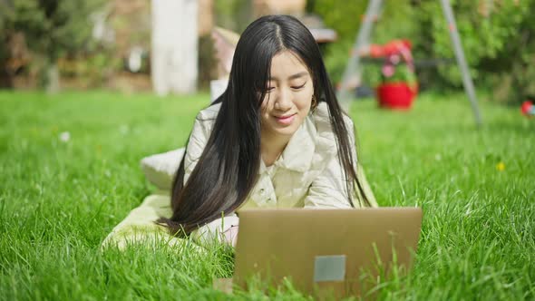 Charming Smiling Millennial Lying on Blanket in Spring Summer Garden Typing on Laptop Keyboard