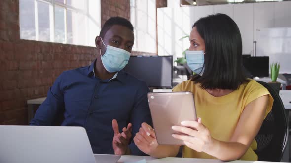 Diverse male and female business colleagues wearing face masks sitting at desk using tablet