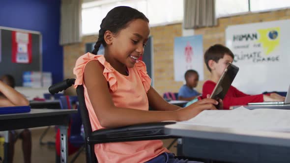 Mixed race schoolgirl in wheelchair, sitting in classroom using tablet, colleagues in background