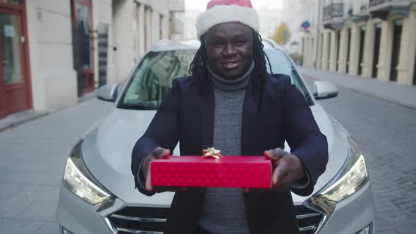 Young Black Man with Santa Hat Giving Red Wrapped Christmas Gift in Front of His Car
