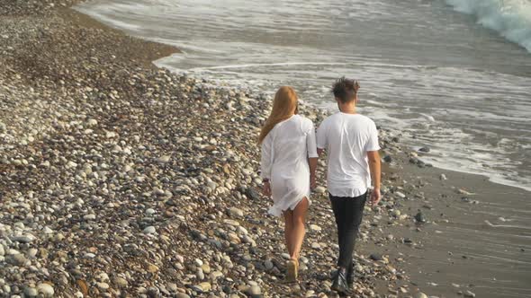 Happy Couple in White Dress Walk Along Rocky Seashore Holding Hands