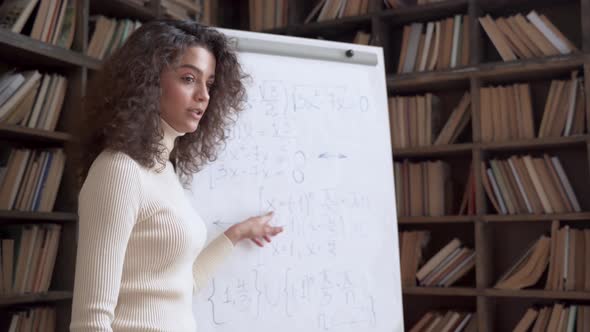 Smiling Hispanic Young Woman Math Teacher Giving Class Standing at Whiteboard