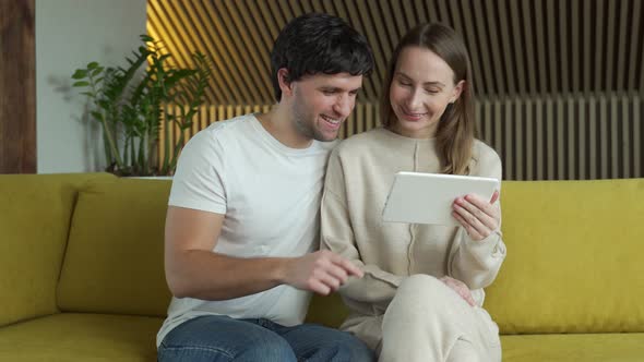 Young Couple Watching Media Content Online in a Tablet Sitting on a Sofa at Home